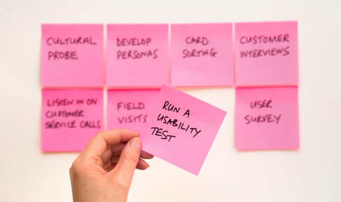 Fotografia di mano che tiene un post-it con la scritta user experience. Sfondo con post-it attaccati