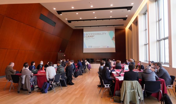 Fotografia dell'evento: sala con quattro tavoli attorno cui sono disposte persone, in fondo alla sala proiettata una slide con scritta Accessibility Camp