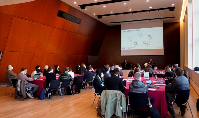 Fotografia dell'evento: sala con quattro tavoli attorno cui sono disposte persone, in fondo alla sala proiettata una slide con scritta Value chain